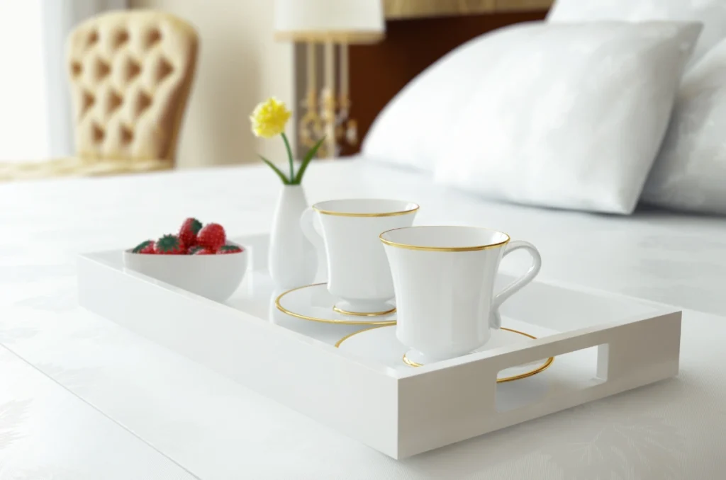 Elegant white tray with gold-rimmed coffee cups, a bowl of strawberries, and a vase with a yellow flower, on a bed in a luxury hotel room