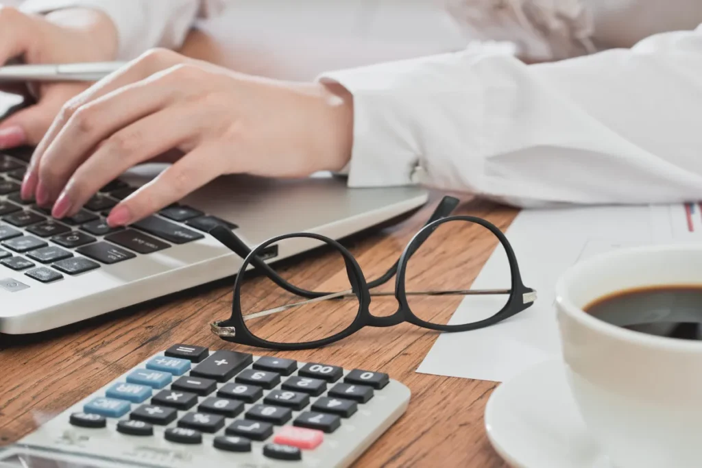 A person's hands are typing on a laptop keyboard. In the foreground, a pair of round-framed glasses rests next to a calculator and a white coffee cup filled with black coffee on a wooden desk. Papers are partially visible under the glasses.
