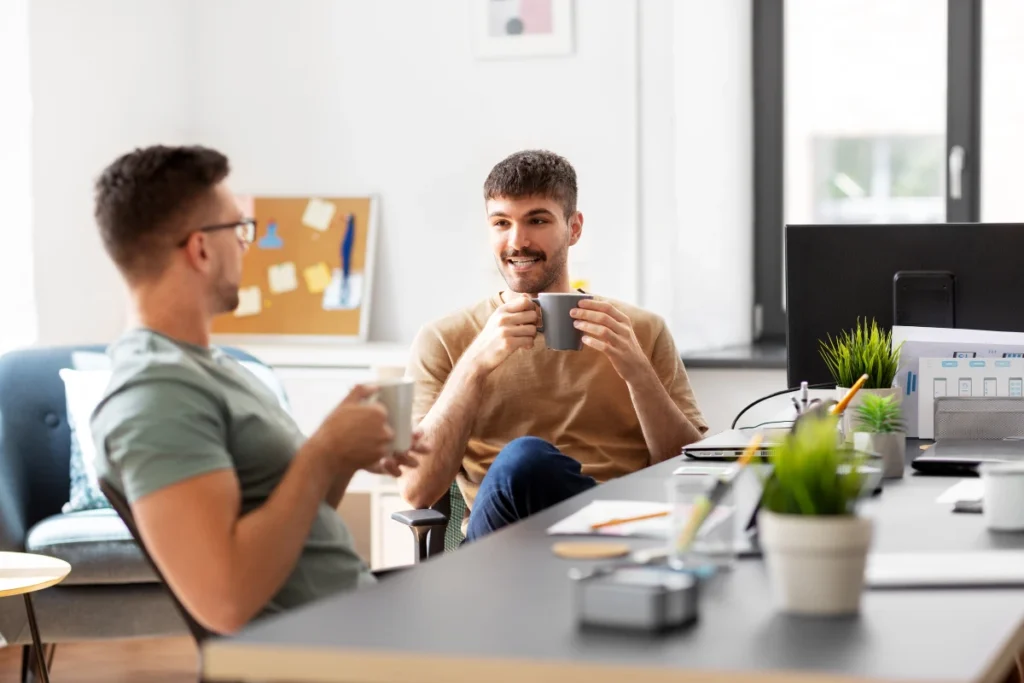 Two men in a casual office setting having a conversation with mugs in their hands. The focus is on the smiling man in the foreground holding a grey mug. In the background, office supplies and a cork board with notes are visible.